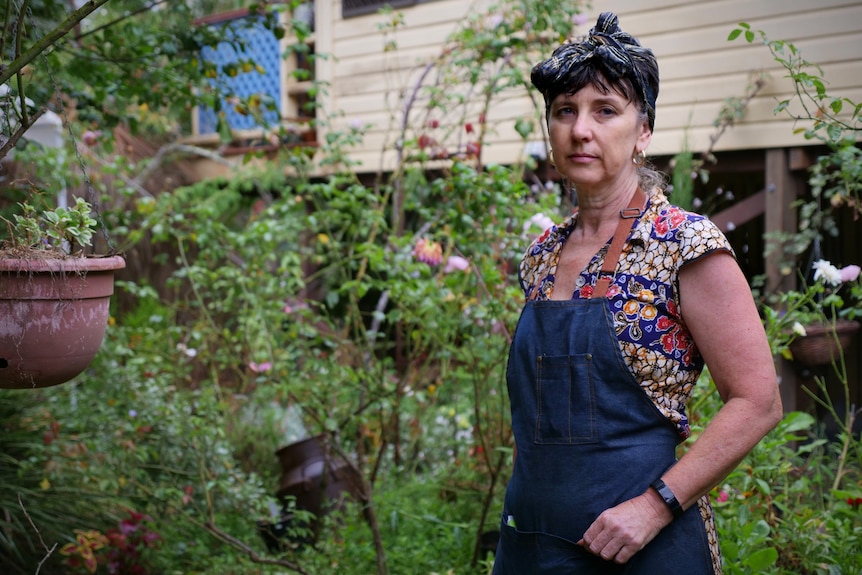 A woman in a headscarf and blue apron stands in the garden outside a raised house.