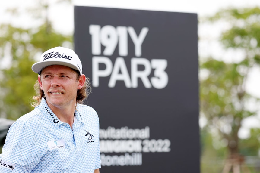 A man with a mullet looks to the side in front of a sign saying "par three".