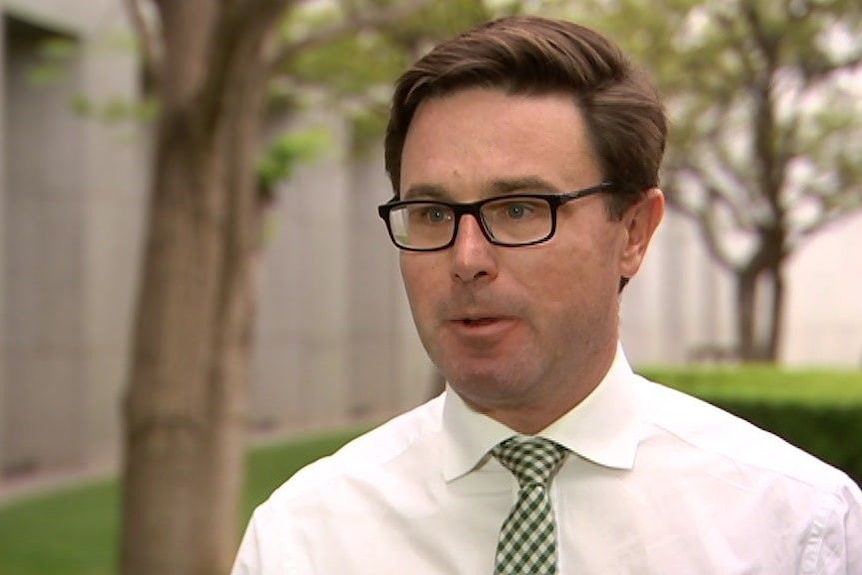 A man in a white shirt, wearing glasses, speaks to the media in an outdoor setting.