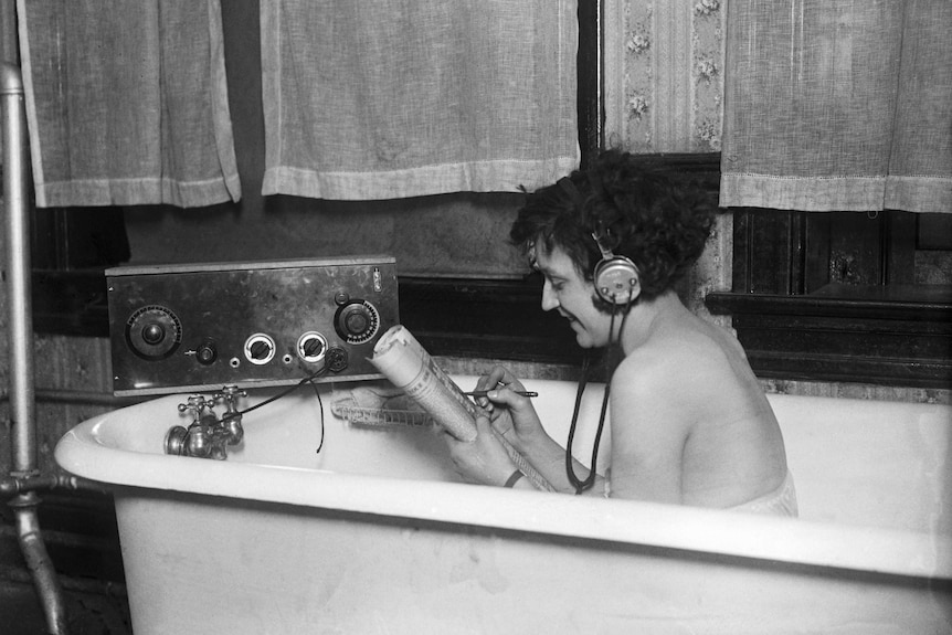 Black and white photo of a smiling woman in the bath listening to a transistor radio, holding the crossword section of a paper.