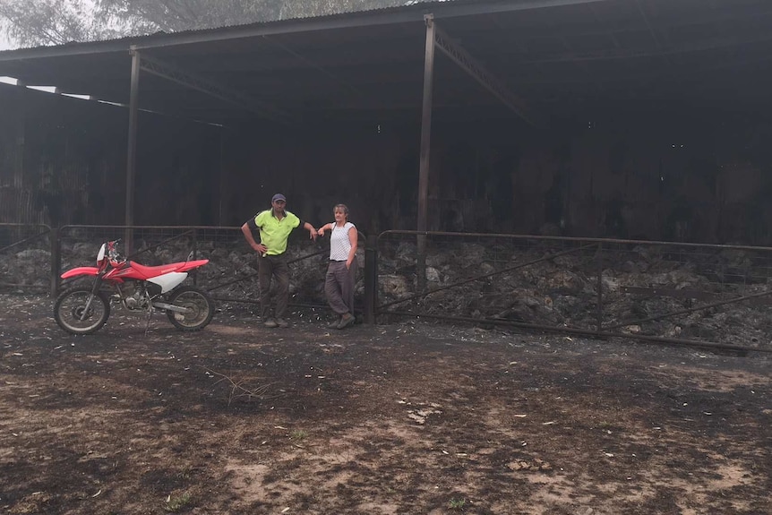 A couple lean on a metal fence with a burnt out farm house behind them.