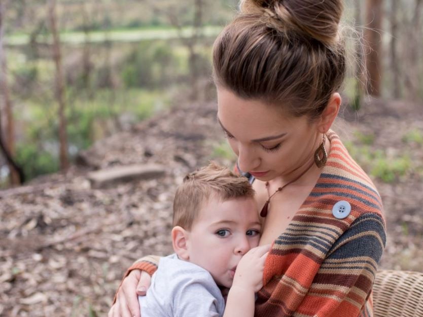 A woman sits on chair breastfeeding toddler son.