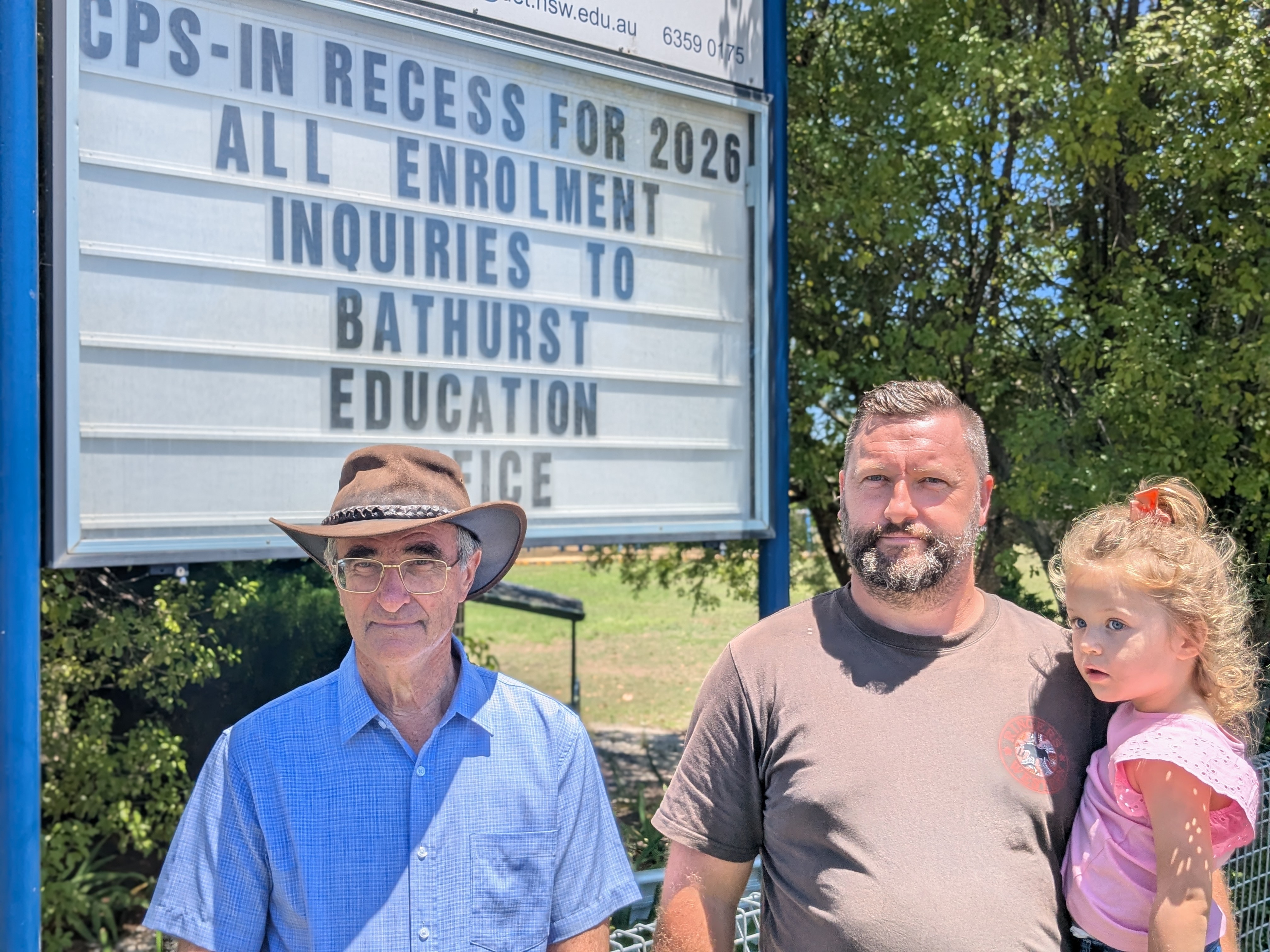 Dos hombres están parados bajo un cartel escolar, uno de ellos sostiene a una niña.