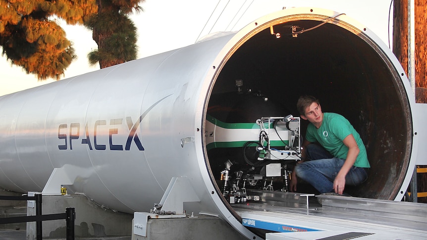 A man kneels over a capsule-shaped metal pod mounted on a track that runs into a large tube.