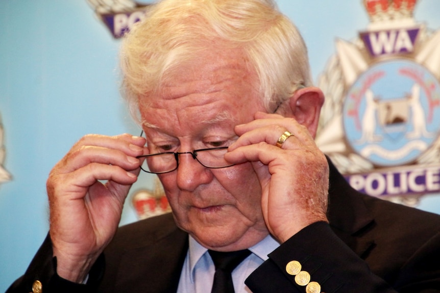 A close-up photo of Denis Glennon adjusting his reading glasses in front of a police backdrop.