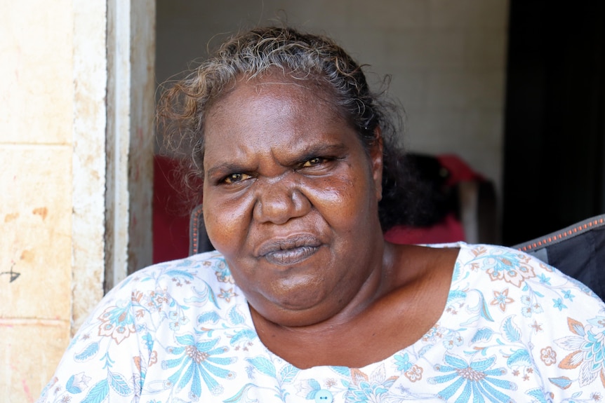 A woman sitting just outside a doorway, looks into the camera with a serious expression on her face.