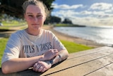 A young woman wearing a t-shirt sits at a bench near a beach