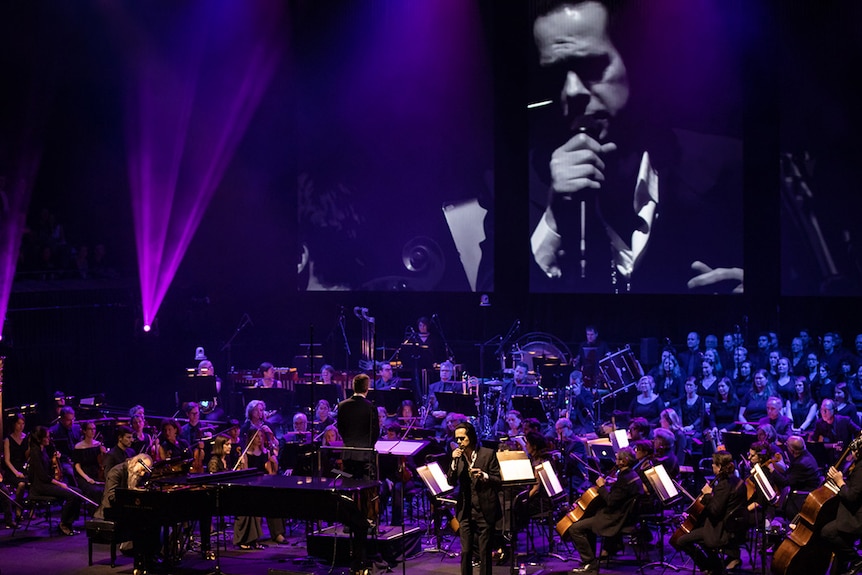 Nick Cave sings in front of an orchestra and choir, his face projected on the screen behind him