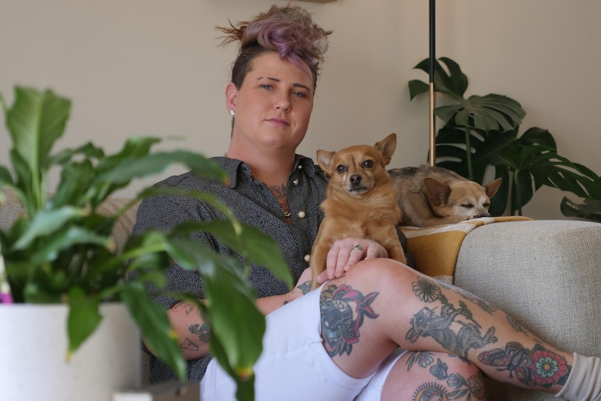 a woman with short pink hair wearing a black shirt sits on a couch with two small dogs.