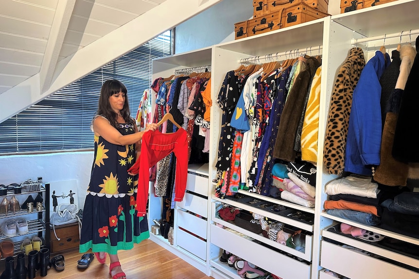 Wide shot of woman looking at red top on hanger in front of large open wardrobe of clothes