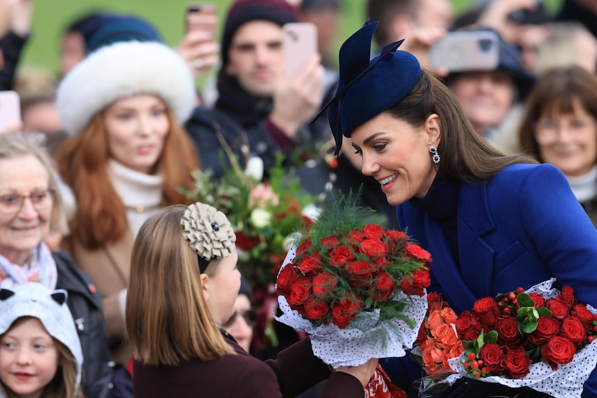 Catherine, wearing a sapphire blue coat and matching hat, collects red roses from members of the public