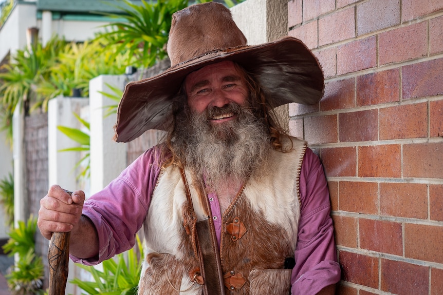 A man with long tawny hair, grey beard and a wizard hat smiles while wearing a skin and fur vest.