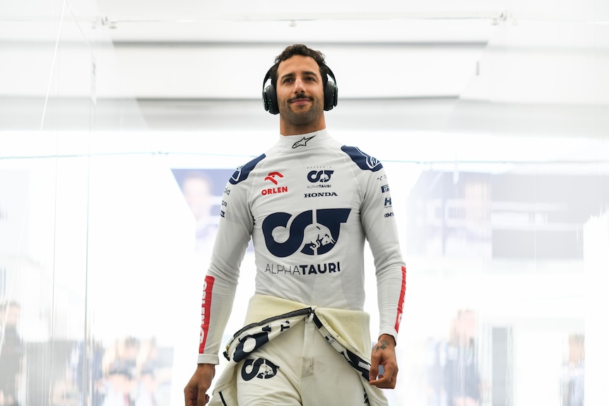 An F1 driver walking through his pit garage, racing suit around his waist