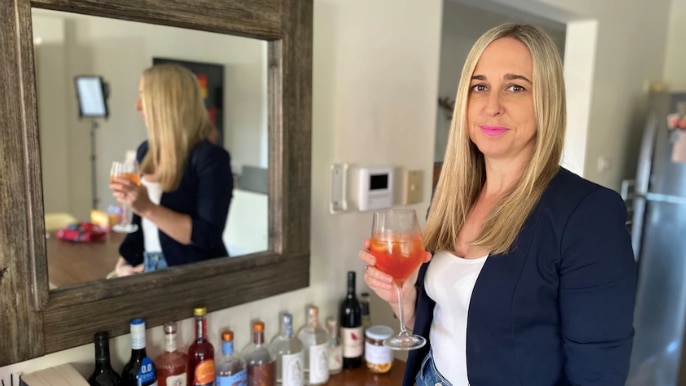 A blonde woman in a navy blazer stands in front of a mirror, holding a wine glass of liquid.