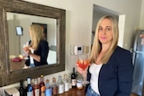 A blonde woman in a navy blazer stands in front of a mirror, holding a wine glass of liquid.