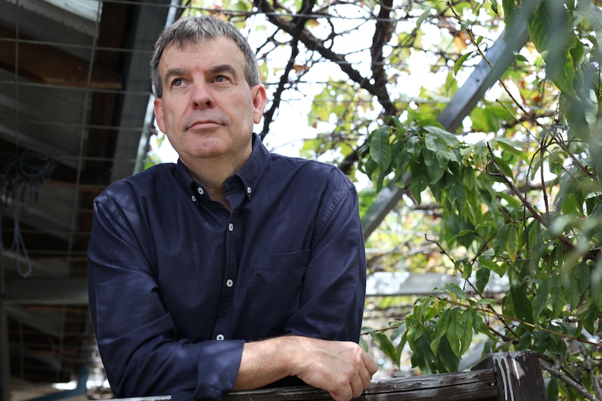 A medium shot of Dave wearing a dark blue shirt, leaning on a wooden post in a backyard, looking into distance
