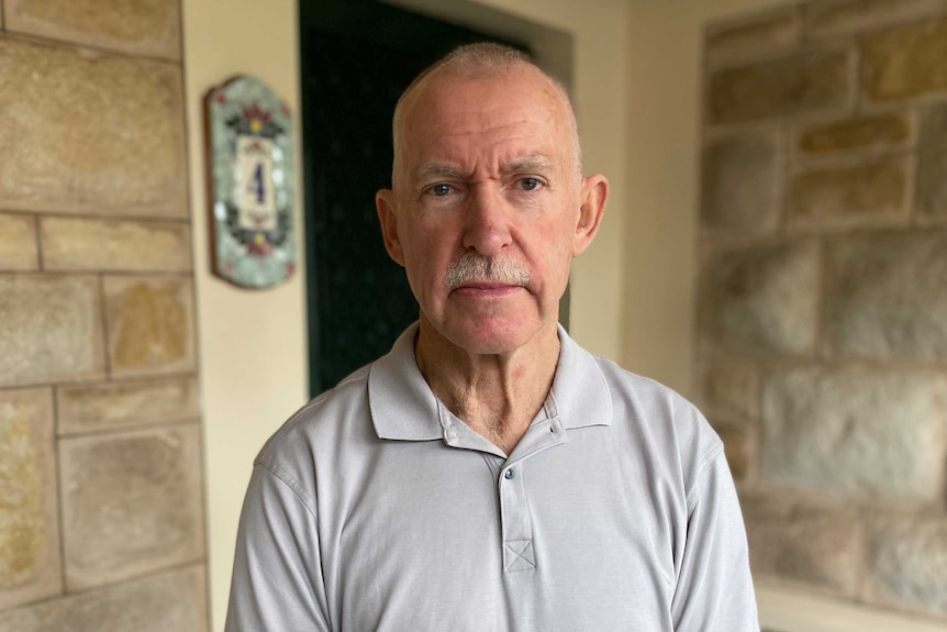 A mostly bald man standing on a verandah with sandstone walls behind him