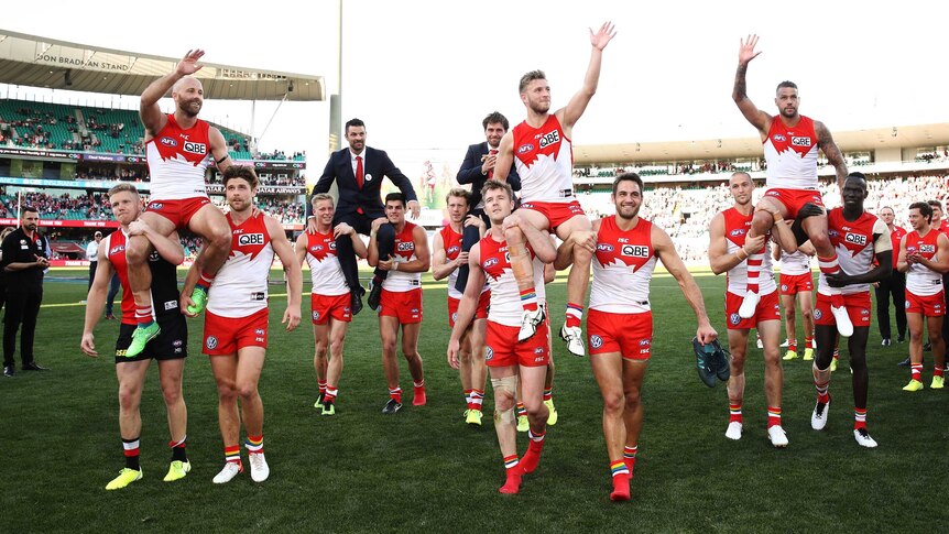 Four male AFL players are chaired off the field by their teammates as they wave to the crowd.