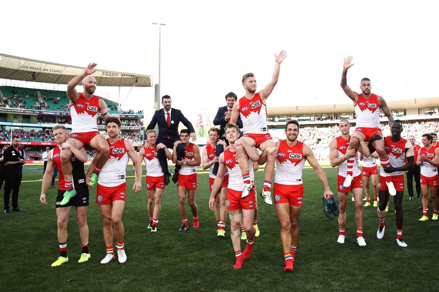 Four male AFL players are chaired off the field by their teammates as they wave to the crowd.