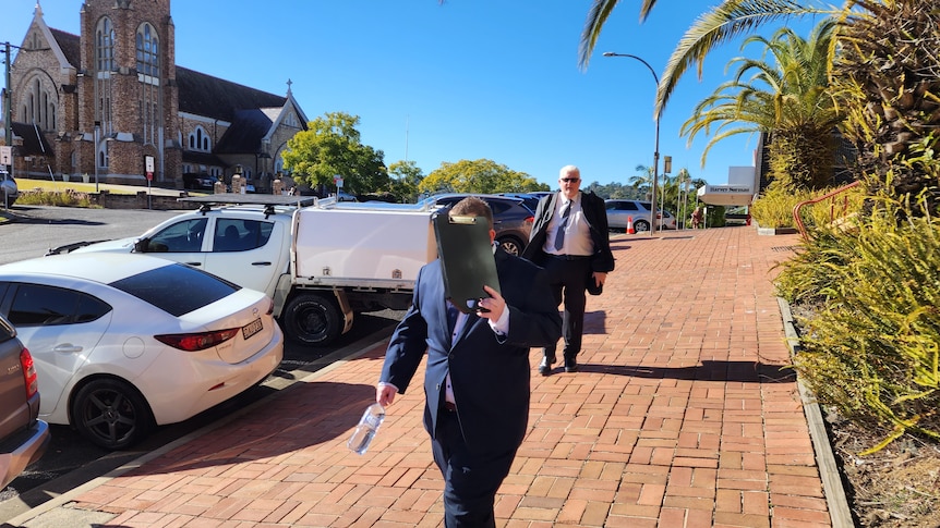 Two men in suits walk down a footpath, one in front of the other. The one in front is shielding his face with a clipboard.