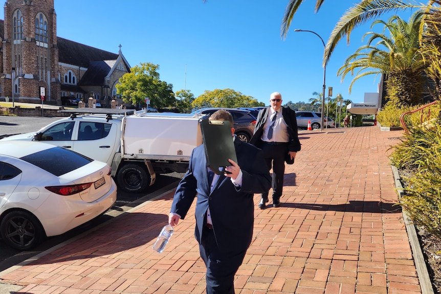 Two men in suits walk down a footpath, one in front of the other. The one in front is shielding his face with a clipboard.