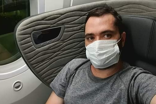 A man with brown hair and eyes and wearing a mask sits on a plane seat next to a window.