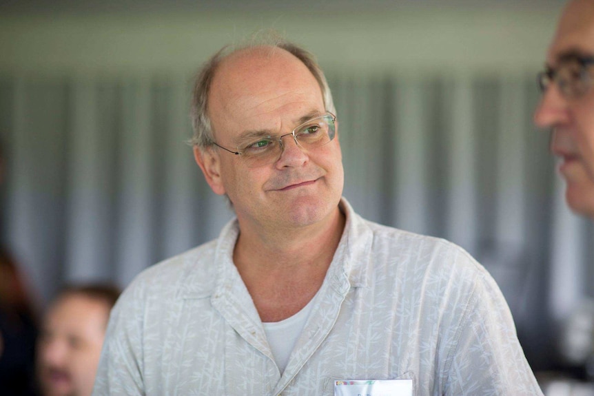 An image of an aging man wearing glasses, with brown eyes, smiling at someone out of shot.