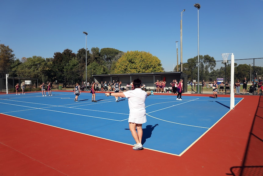 Young women wearing uniforms move about a brightly coloured outdoor netball court, watched by an umpire wearing white