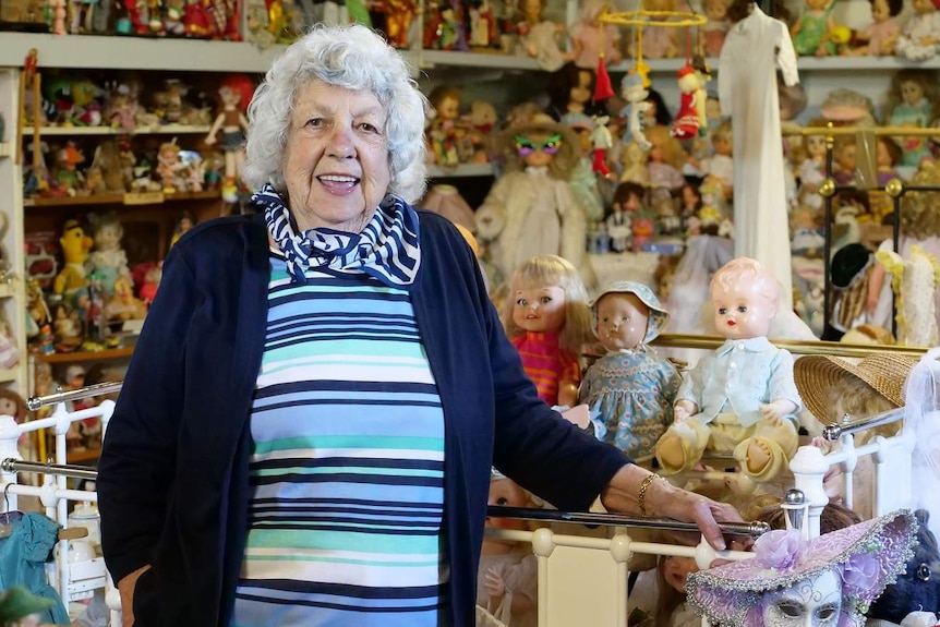 A smiling, elderly woman with short grey hair surrounded by dolls of all shapes, sizes and styles.