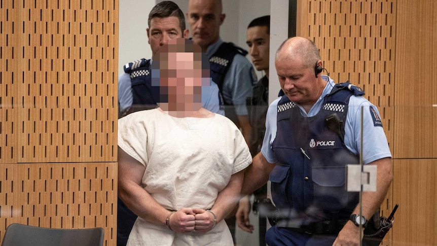 Brenton Tarrant, face pixelated and wearing a white gown, is lead into the dock of the Christchurch District Court.