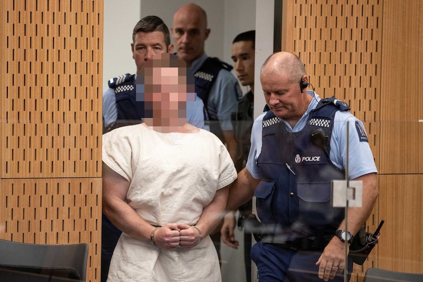 Brenton Tarrant, face pixelated and wearing a white gown, is lead into the dock of the Christchurch District Court.