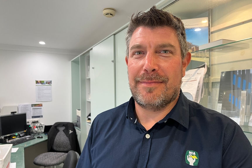 Daniel Pleaner look into camera lens, standing in an office, wearing a HIA shirt.