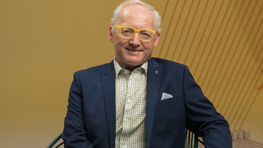 Toby Walsh, a man wearing yellow framed spectacles and a navy blazer, poses for a photo in front of an ochre coloured background