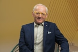 Toby Walsh, a man wearing yellow framed spectacles and a navy blazer, poses for a photo in front of an ochre coloured background