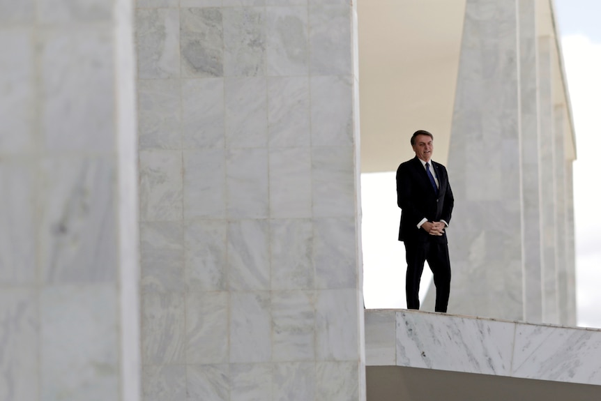 You view a small Jair Bolsonaro in a black suit as he stands on the ramp of the Planalto Palace, dwarfed by the structure.
