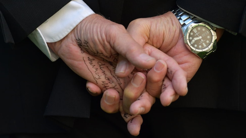 close up shot of man's hands in formal attire and watch clasped tightly together behind back. There is writing on the man's palm