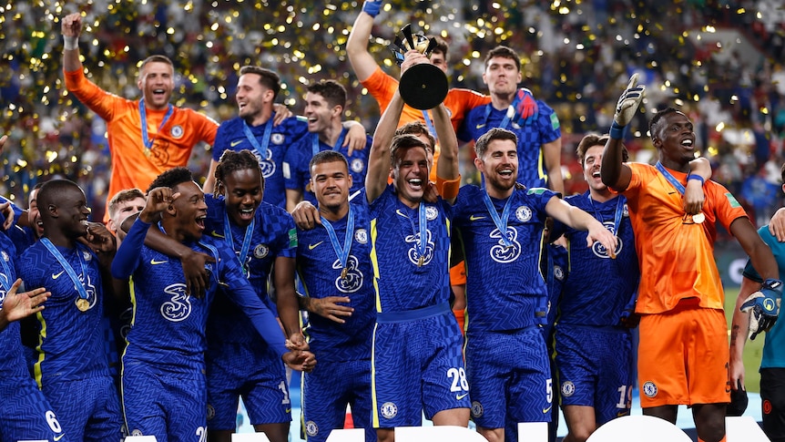 A soccer team wearing blue kits celebrates winning a competition, lifting a trophy and wearing winners medals
