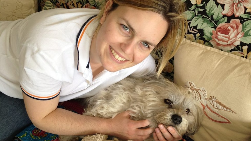 A woman smiles with her small white dog on a rug on the floor.