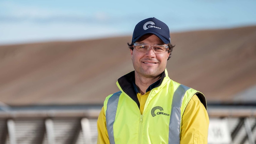 Man wearing a high-vis vest and shirt and blue baseball cap and safety glasses, with wheat stack blurred in background