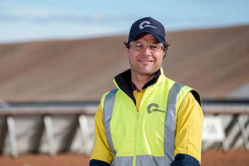 Man wearing a high-vis vest and shirt and blue baseball cap and safety glasses, with wheat stack blurred in background