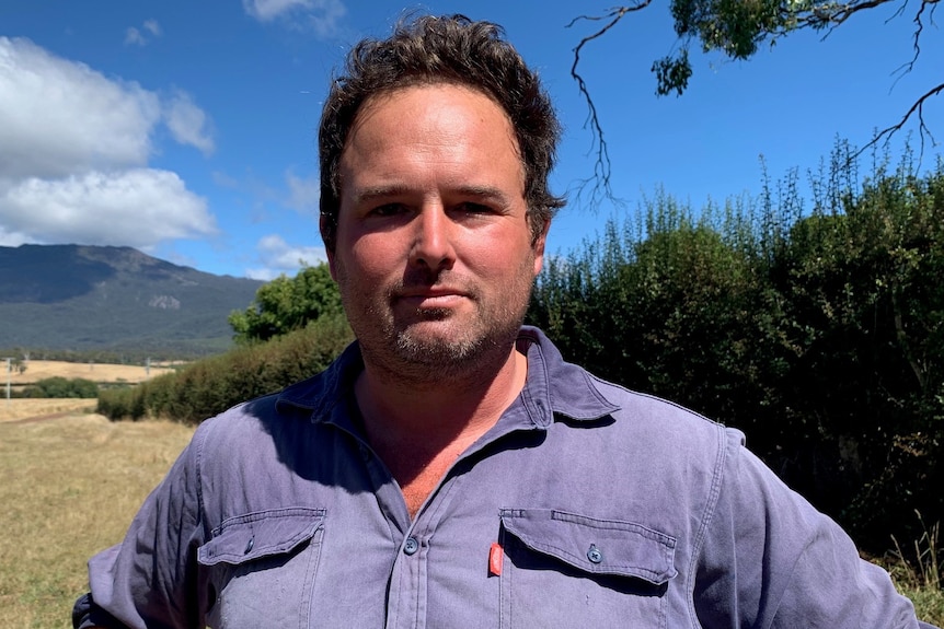 A man in a blue shirt looks seriously at the camera, farmland rolling away in the distance behind him.