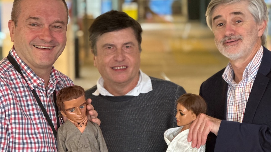 three men pose together, two holding puppets that look like humans