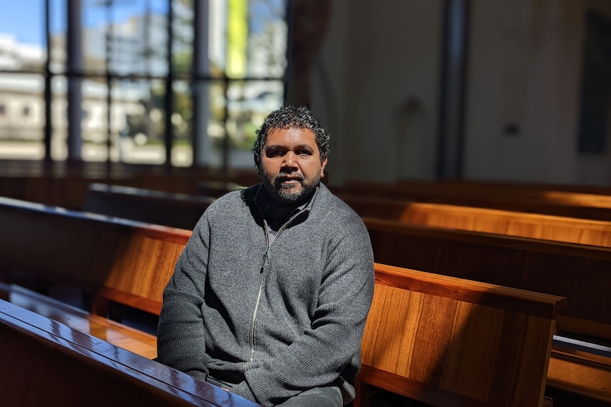 An Indigenous man in a grey jacket sits in a patch of sunlight on church pews inside a building.