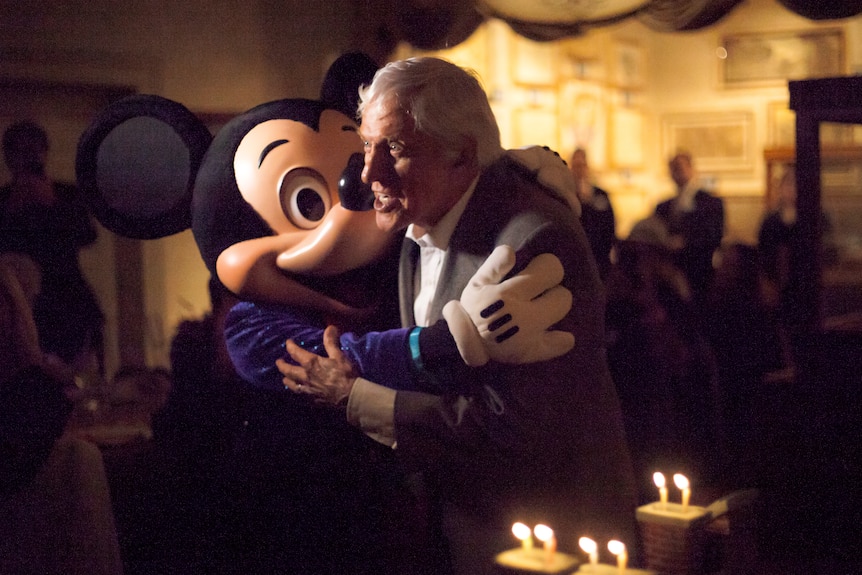 An elderly man embraces someone in a Micky Mouse costume.