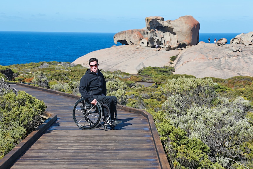 A man in a wheelchair on a wooden walkway smiles at the camera with a stone structure and the ocean behind him