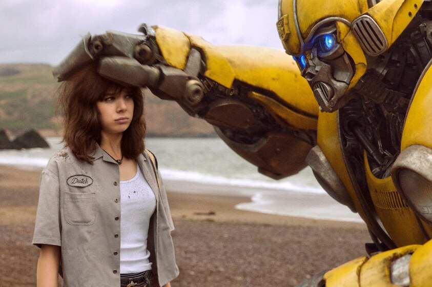On an overcast day at the beach a towering yellow robot pats the head of an unimpressed young woman with brown hair.