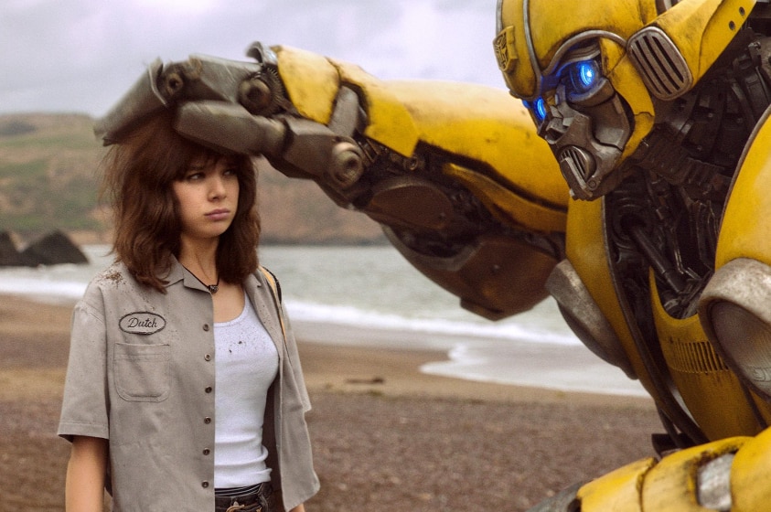 On an overcast day at the beach a towering yellow robot pats the head of an unimpressed young woman with brown hair.