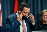 Mark McGowan puts a black mask on his face, in a room with a blue curtain and australian flags behind him.