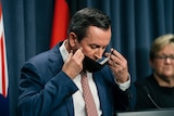 Mark McGowan puts a black mask on his face, in a room with a blue curtain and australian flags behind him.