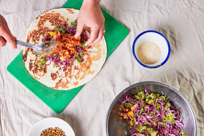 A cook spoons the chicken filling into the burrito with a bowl of cabbage, kale and corn slaw, for a family dinner recipe.
